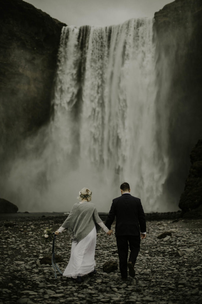 bride and groom holding hands walking towards waterfall