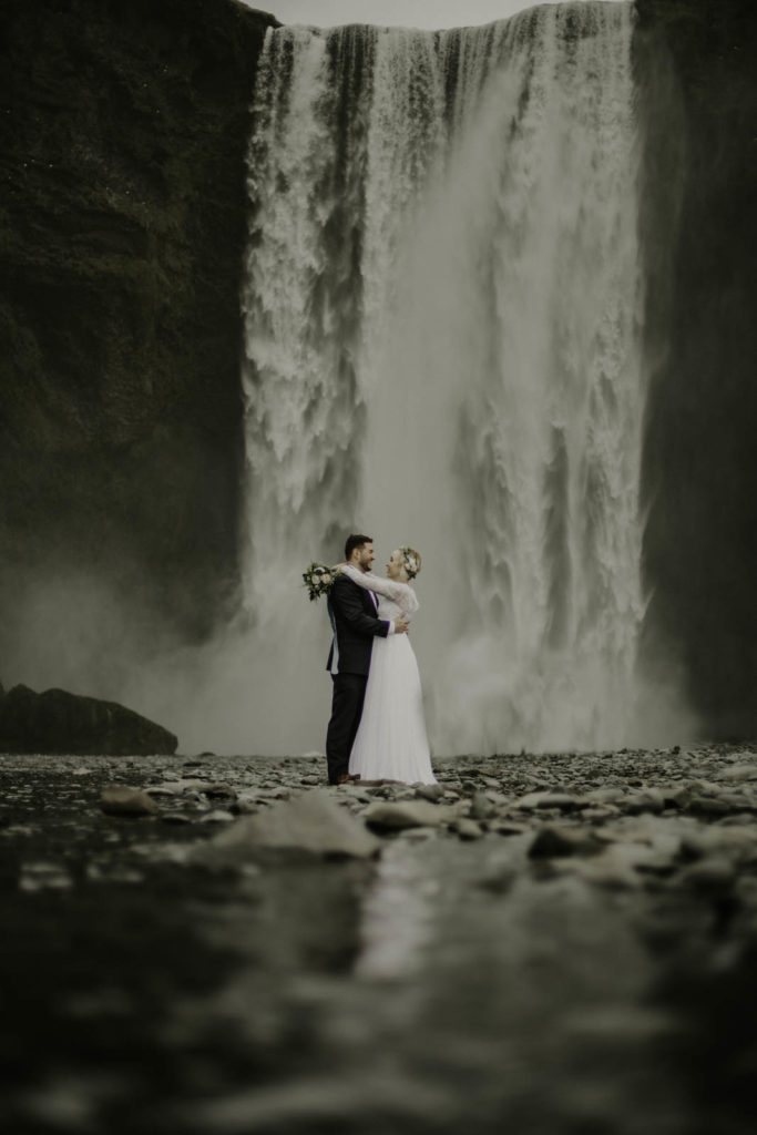bride and groom standing in river next to waterfall