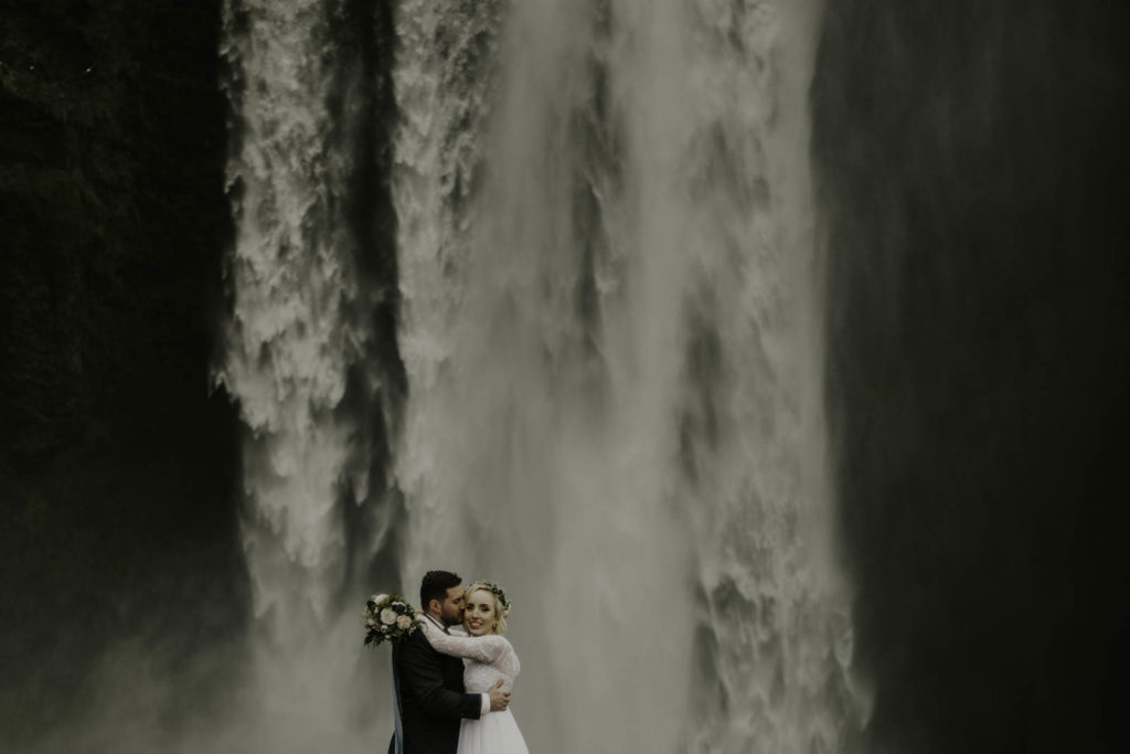 groom kissing bride on cheek in front of waterfall