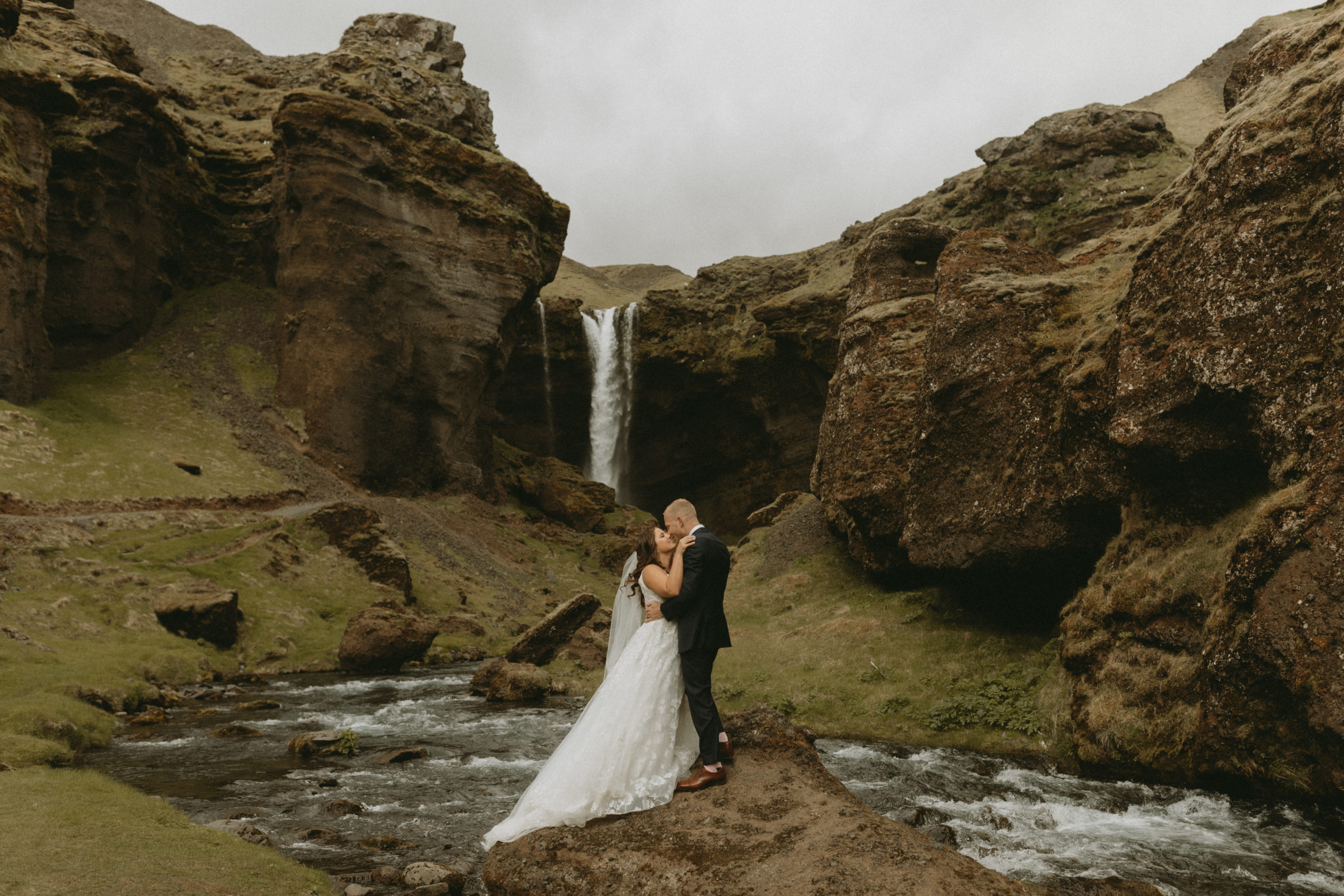 Bride and groom standing on rock in river with an iceland waterfall behind them.
