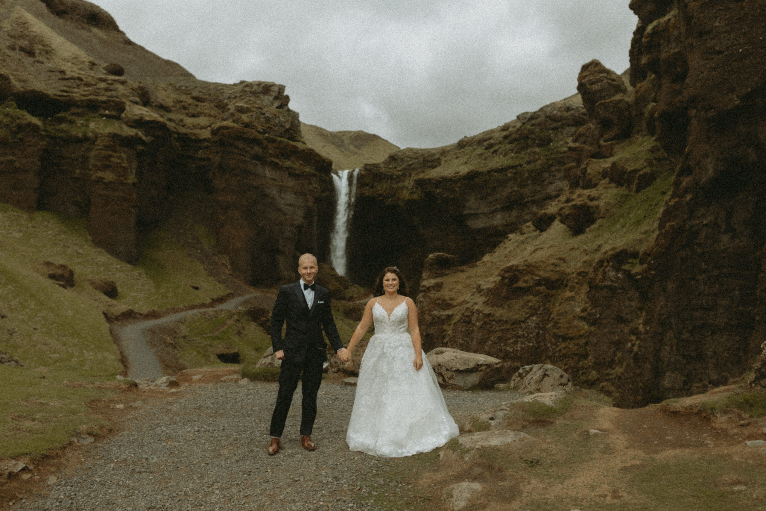 Bride and groom standing in Iceland canyon on wedding day with a waterfall behind them.