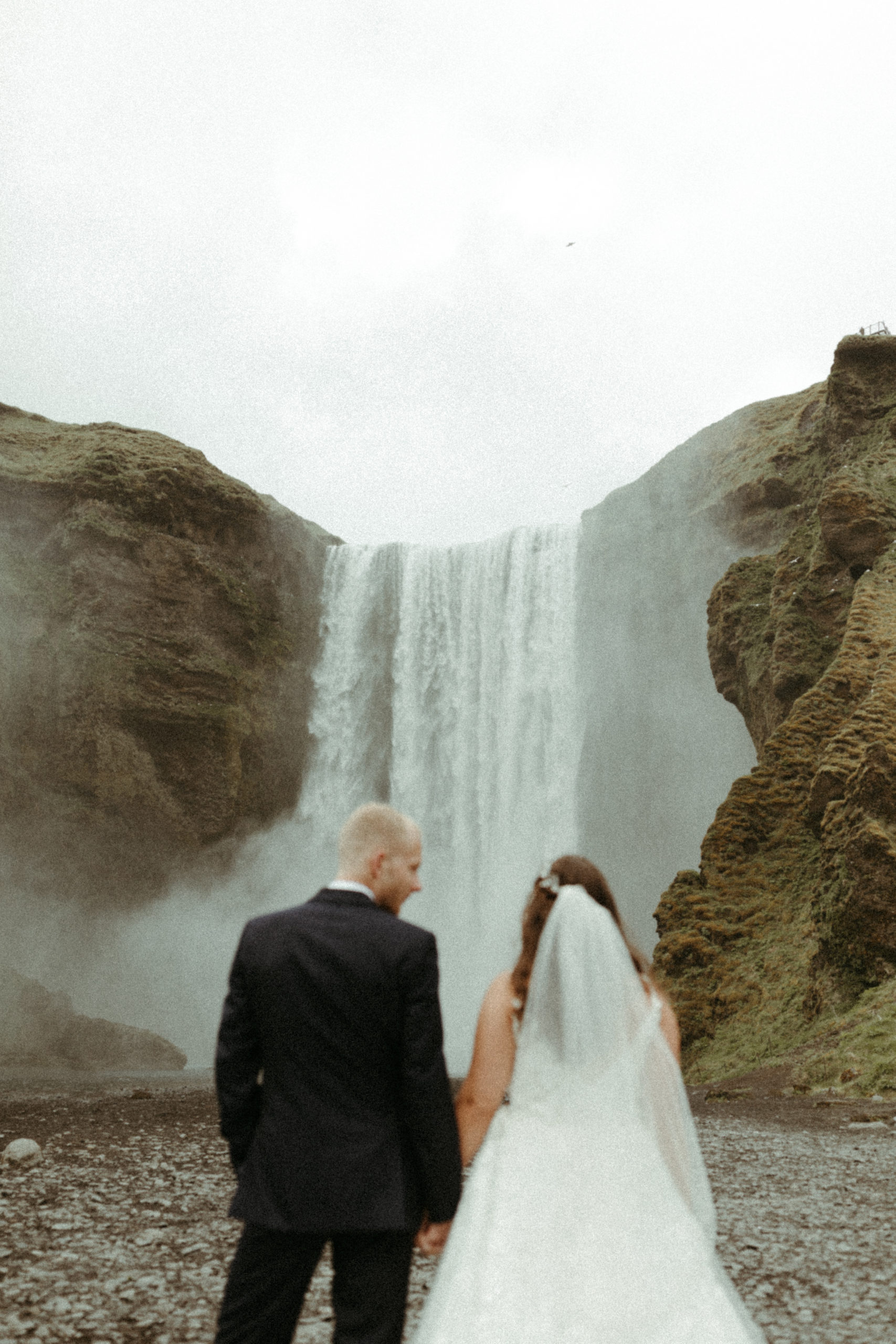 Bride and groom standing in front of Skogafoss waterfall in Iceland on wedding day.