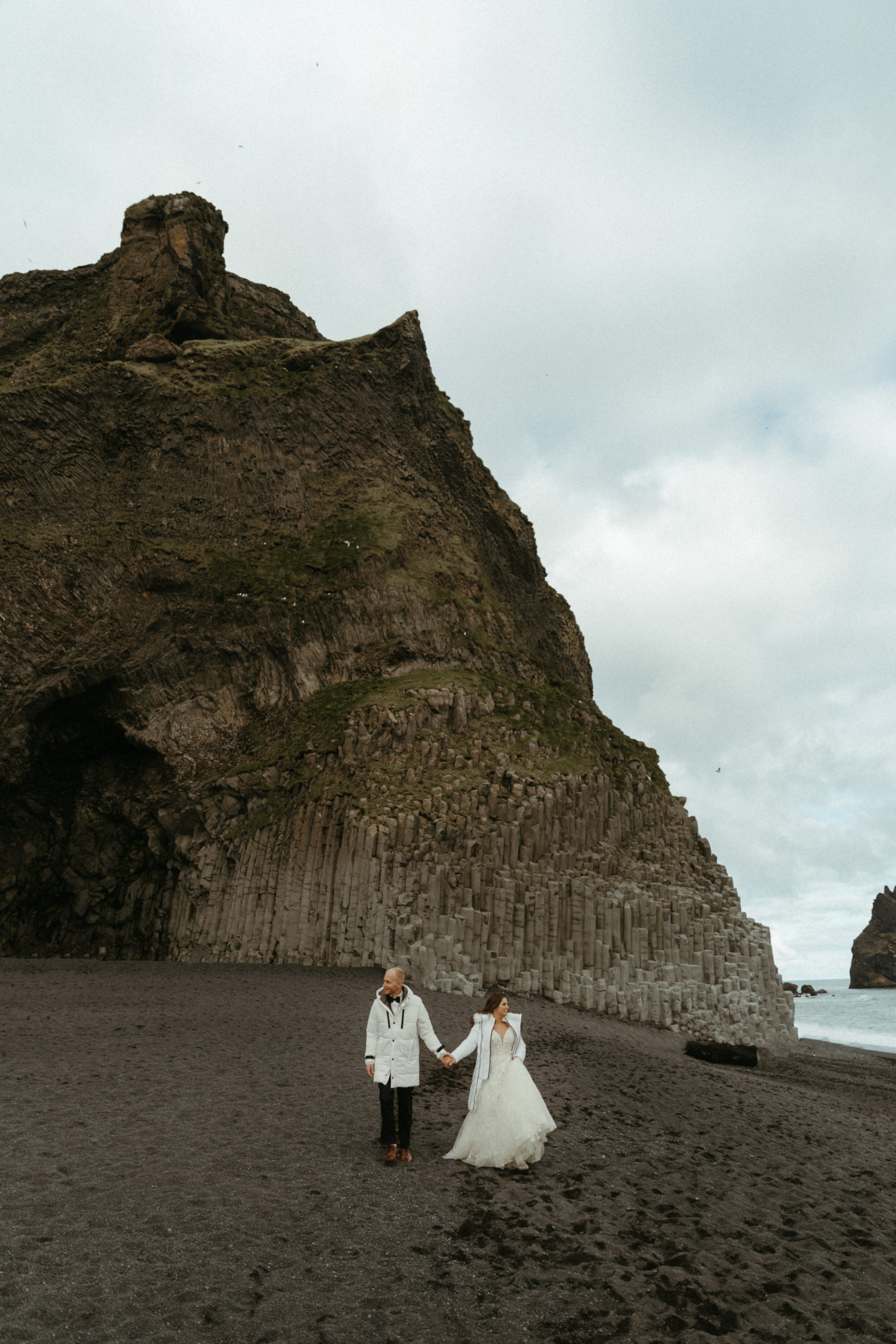 Bride and Groom walking down black sand beach in Southern Iceland on wedding day. 