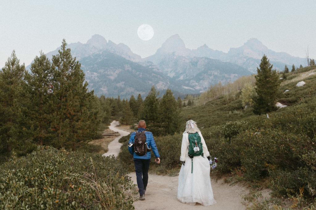 Bride and groom hiking towards mountains with a full moon behind them.