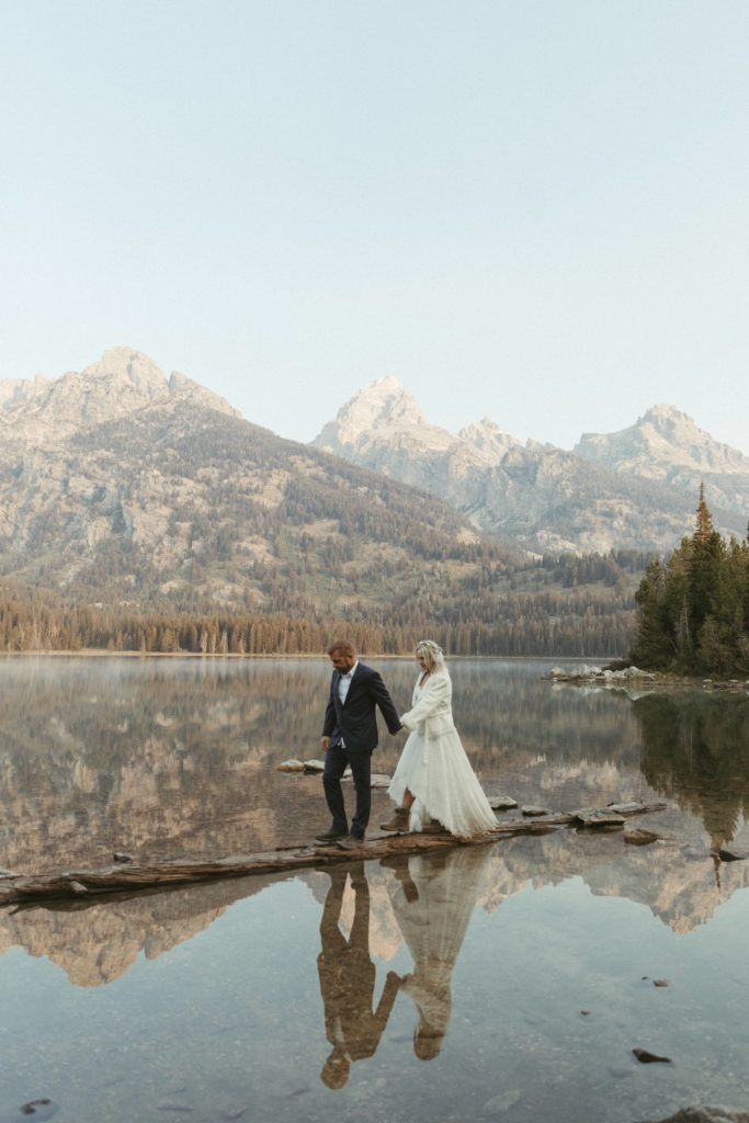 Bride and Groom walking across a log in a lake with the Grand Teton mountains behind them.