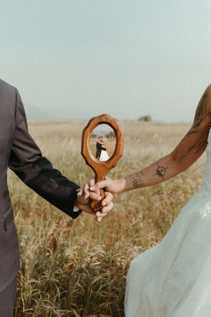 Bride and groom holding mirror together in a field with the mountains behind them.