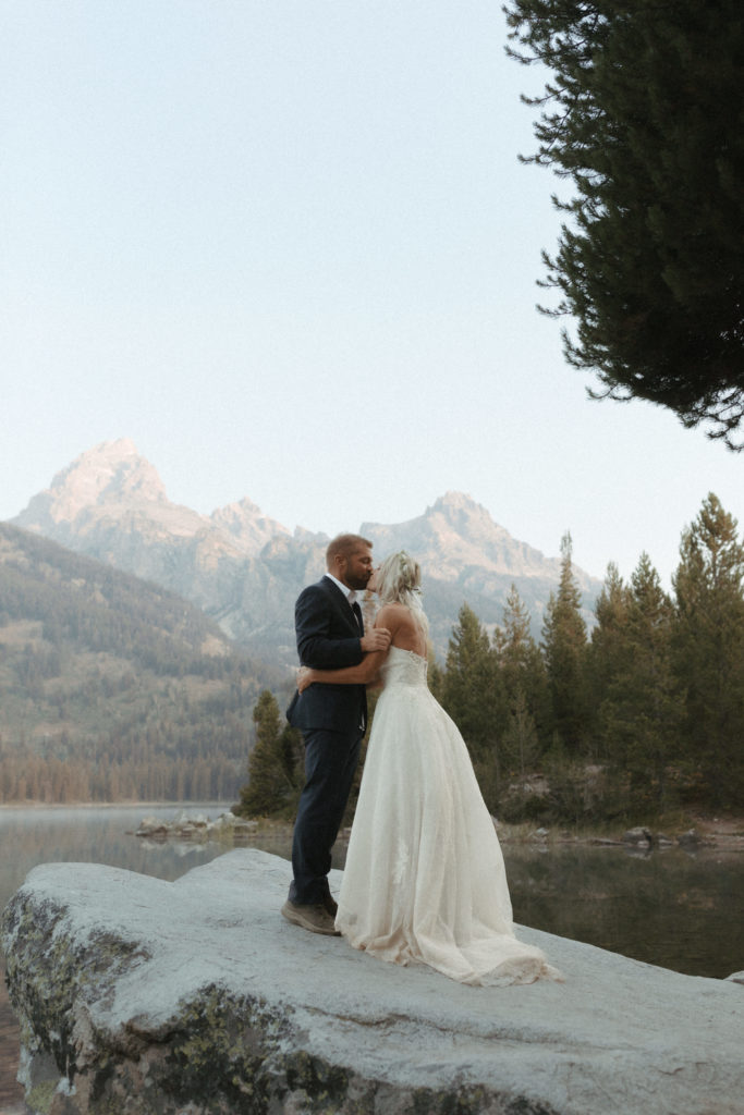 Bride and groom kissing on rock by lake in the mountains.