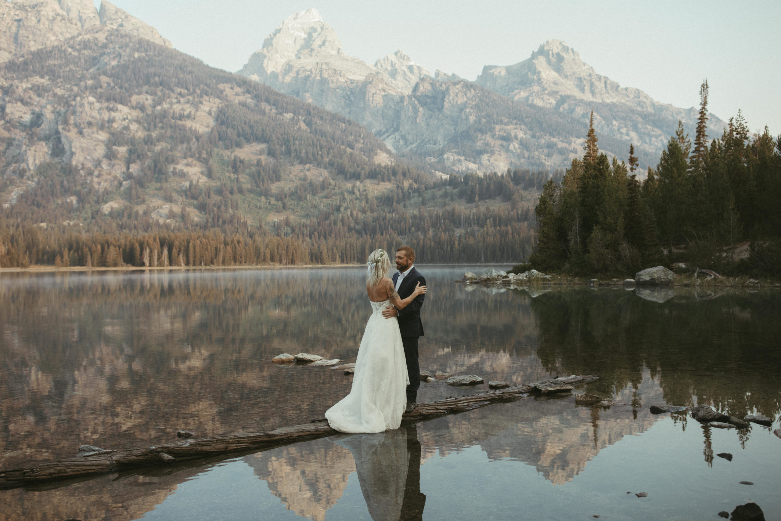 Bride and Groom standing on a log in a lake with the Grand Teton mountains behind them.