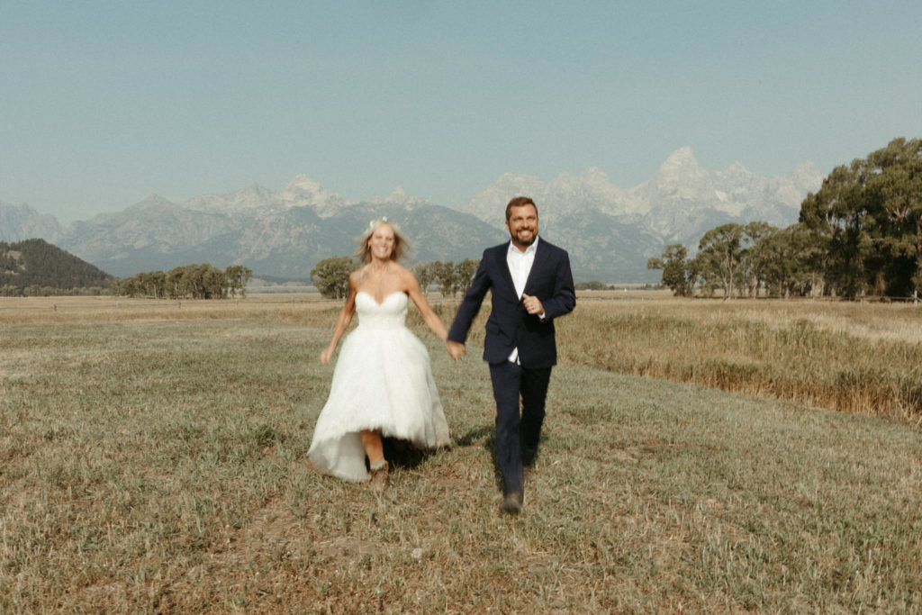 Bride and groom running through golden fields with mountains behind them.
