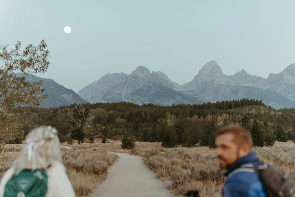 Bride and groom hiking on trail with a full moon over the mountains.